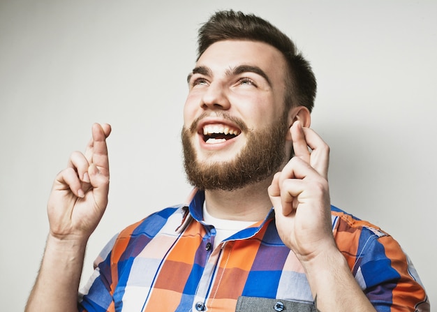 Life style and people concept:Waiting for special moment. Portrait of young bearded man in shirt keeping fingers while standing against white background. Hipster style and positive emotions.