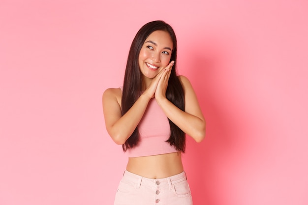 Lifestyle, beauty and women concept. Portrait of dreamy and cute asian girl in summer clothes, smiling and looking up while lying on palms as if going to bed, standing pink wall