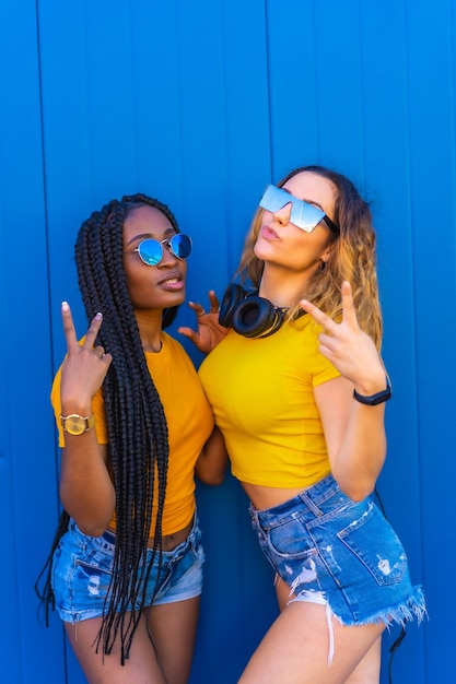 Lifestyle, black girl and blonde Caucasian girl in yellow dresses. Very happy and smiling with music headphones and sunglasses