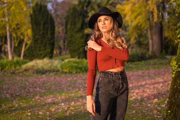 Lifestyle blonde Caucasian girl in a red sweater and black hat enjoying nature in a park with trees portrait of the young woman enjoying the autumn sun