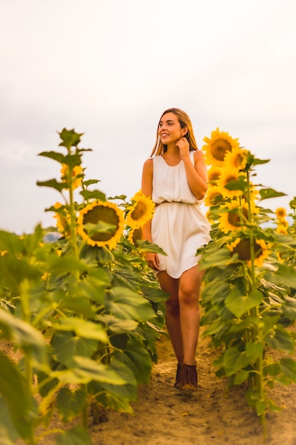 Lifestyle, blonde girl in a white dress and a belt walking among sunflowers, looking to the left