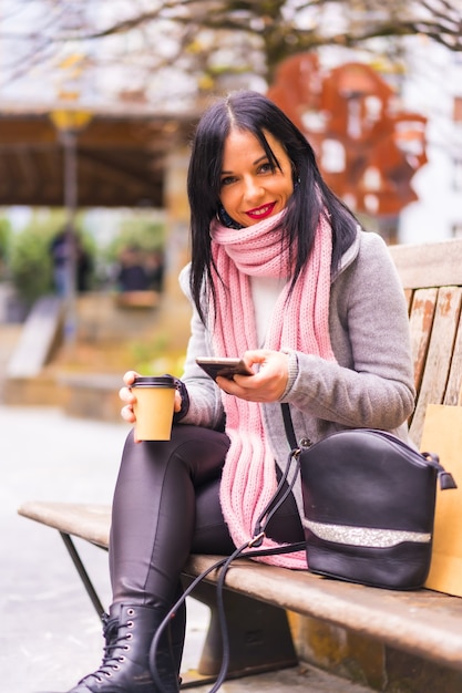 Lifestyle, a Caucasian brunette girl shopping in the city with paper bags and a take away coffe, sitting sending a message with the phone