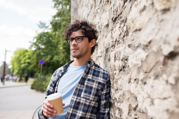 lifestyle, drinks and people concept - man in eyeglasses drinking coffee from disposable paper cup over stone street wall