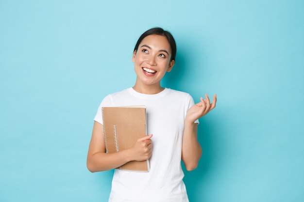 Lifestyle, education and people concept. Happy beautiful asian female student in white t-shirt, girl talking and looking dreamy upper left corner while holding notebooks, light blue wall