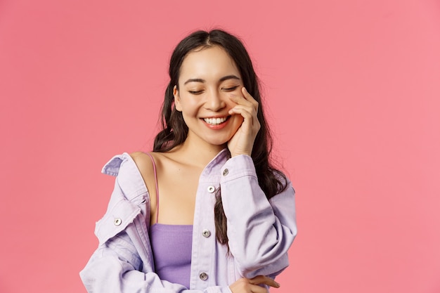 Lifestyle, fashion and beauty concept. Close-up portrait of cheerful, lovely young asian woman close eyes, enjoying day, laughing happily, touch clean skin, pink wall