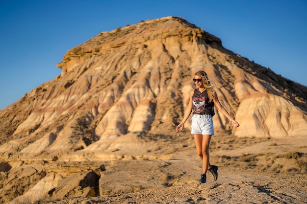 Lifestyle, happy blonde Caucasian girl in black boots, sunglasses and shorts strolling through the Bardenas desert. Spain