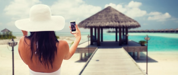 lifestyle, leisure, summer, technology and people concept - smiling young woman or teenage girl in sun hat taking selfie with smartphone over bungalow on beach background