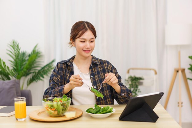 Lifestyle in living room concept Young Asian woman looking at tablet and eating vegetable salad