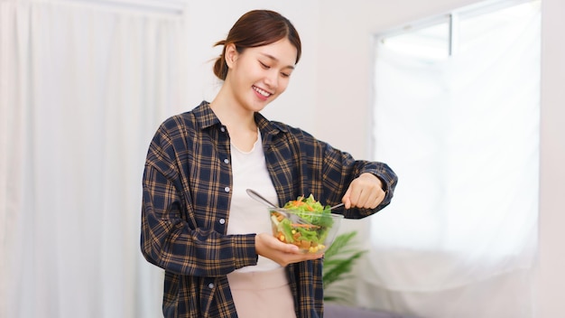 Lifestyle in living room concept Young Asian woman standing to mixing vegetable salad in bowl