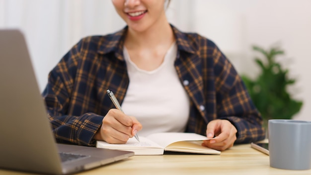 Lifestyle in living room concept Young Asian woman using laptop and taking notes data on notebook