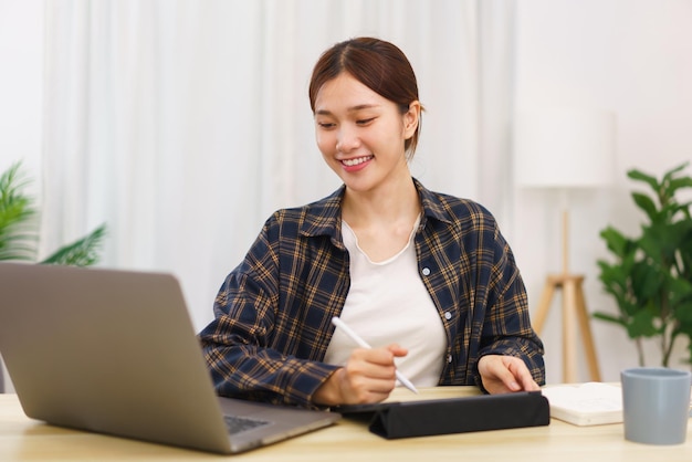 Lifestyle in living room concept Young Asian woman using laptop and taking notes data on tablet