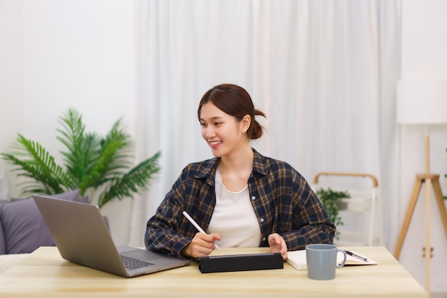 Lifestyle in living room concept Young Asian woman using laptop and taking notes data on tablet