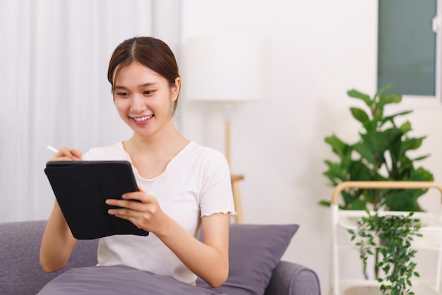 Lifestyle in living room concept Young Asian woman writing data on tablet while sitting on couch