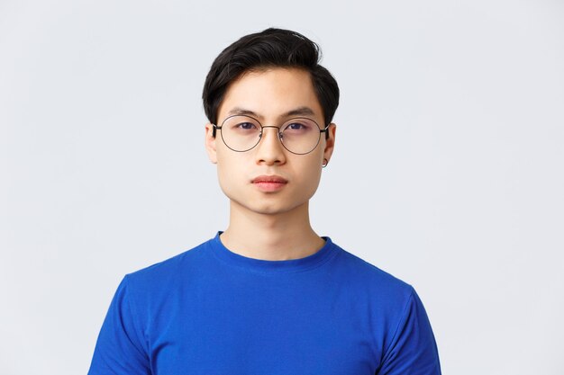 Lifestyle, people emotions and beauty concept. Close-up of asian man in glasses with stylish haircut looking at camera with normal, calm expression, standing grey background