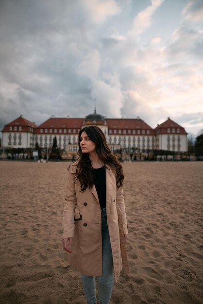 Lifestyle portrait of brunette wears trench coat black shirt and jeans on the ocean coast Girl posing near classical hotel by the seaside during cold time of the year