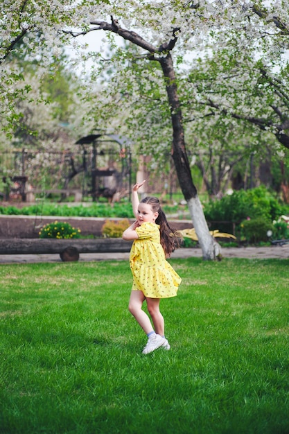 Lifestyle portrait of a cheerful girl in a yellow dress dancing on the green grass in a garden with ...