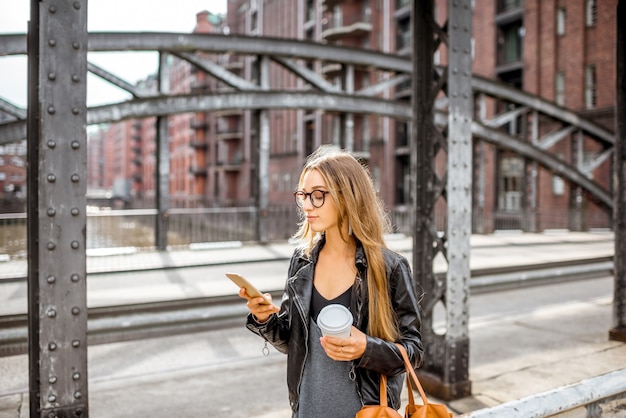 Lifestyle portrait of a stylish business woman with smartphone and coffeecup walking outdoors on the old iron bridge