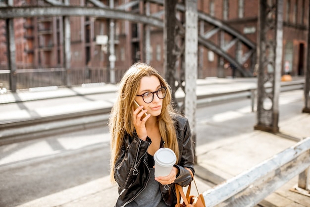 Lifestyle portrait of a stylish businesswoman in leather jacket sitting with smartphone and coffeecup on the old iron bridge