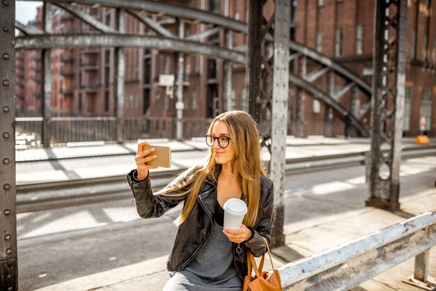 Lifestyle portrait of a stylish businesswoman in leather jacket sitting with smartphone and coffeecup on the old iron bridge