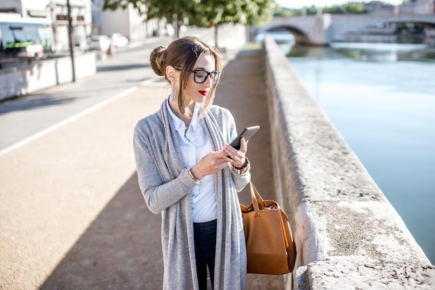 Lifestyle portrait of a stylish businesswoman using phone near the river in Lyon old town during the morning in France