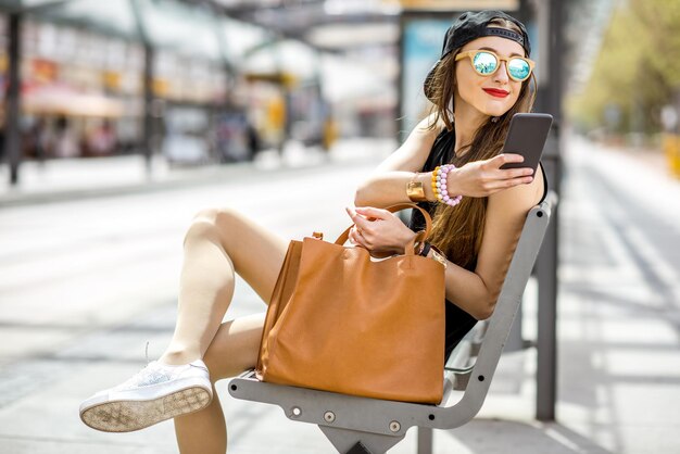 Lifestyle portrait of a stylish woman in black dress and hat sitting with phone on the modern tram stop