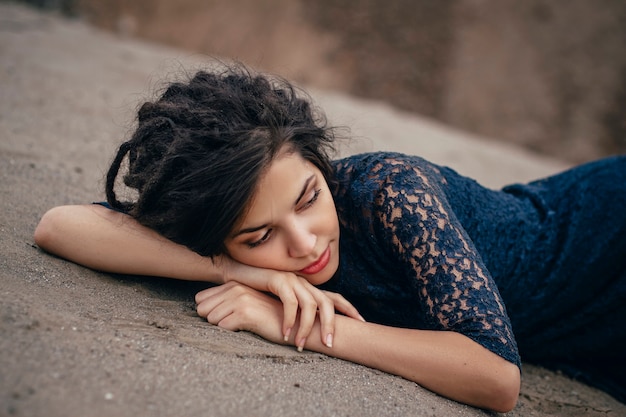 Lifestyle portrait of a woman brunette on background of the lake lying in sand on a cloudy day. Romantic, gentle, mystical, pensive image of a girl. Girl Oriental appearance, dreams of lying on sand