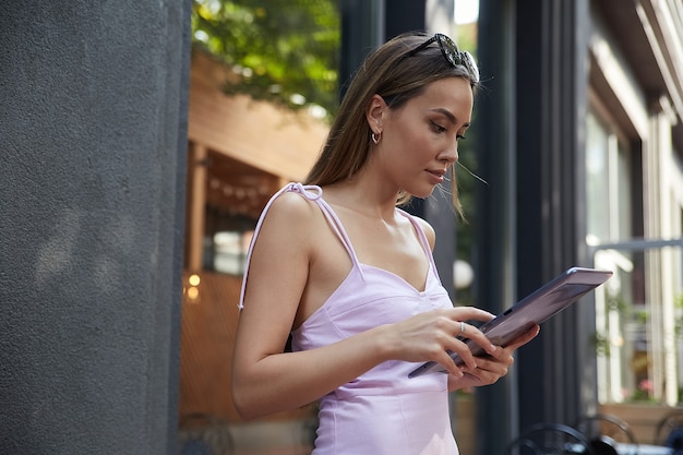 Lifestyle portrait of young asian pretty girl in pink dress using tablet outdoor