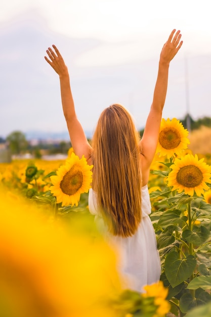 Lifestyle, a pretty caucasian blonde in a field of sunflowers in a white dress, with her arms up