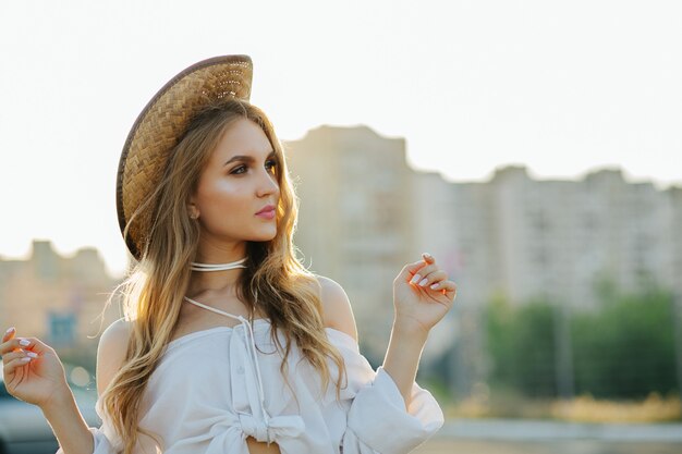 Lifestyle shot of glamor blonde woman wearing trendy apparel and hat posing in sunny evening. Space for text