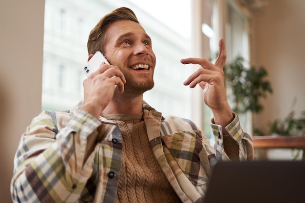 Lifestyle shot of young happy man sitting in cafe with laptop answer phone call talking to friend on