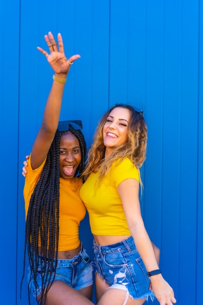 Lifestyle, teenage girl friends enjoying in the photoshoot on a blue wall dressed in yellow t-shirts. Black girl with long braids and blonde Caucasian girl.