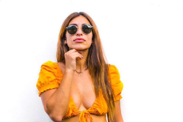 Lifestyle of a young caucasian brunette with orange t-shirt, sunglasses and short pants on a white background, with copy space and sticks. With a sweet and seductive look