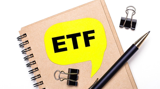 On a light background a brown notebook a black pen and paper clips and a yellow card with the text ETF Exchange Traded Funds