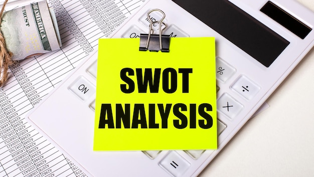 On a light background cash a white calculator and a yellow sticker under a black paper clip with the text SWOT ANALYSIS Business concept