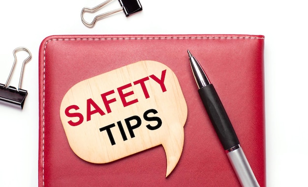 On a light background there are black paper clips a pen a burgundy notepad a wooden board with the text SAFETY TIPS