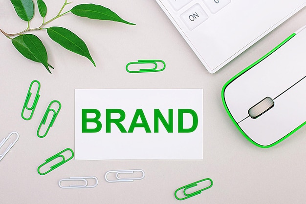 On a light background a white calculator a computer mouse green paper clips a green plant and a white blank sheet with the text BRAND Flat lay