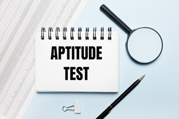 On a light blue background, reports, a magnifying glass, a pen and a notebook with the text APTITUDE TEST. Business concept. Flat lay.