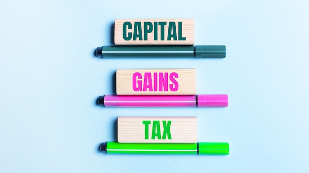 On a light blue background, there are three multi-colored felt-tip pens and wooden blocks with the CAPITAL GAINS TAX