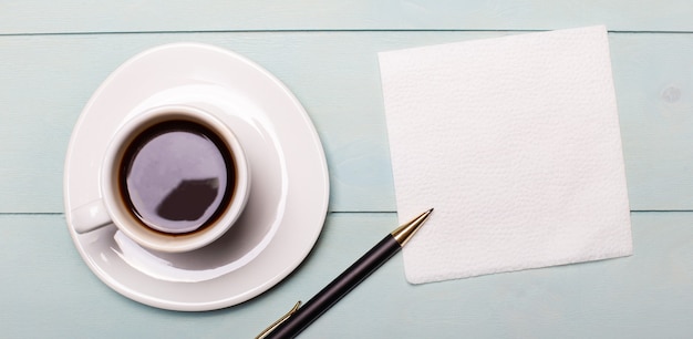 On a light blue wooden background, a white cup of coffee, an empty napkin for notes and a pen. Top view with copy space