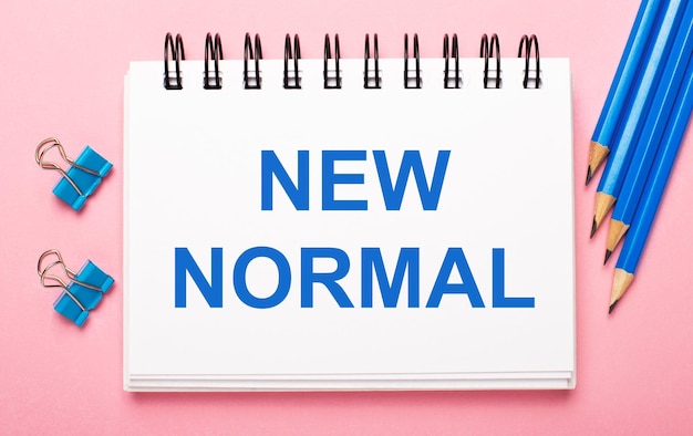 On a light pink background, light blue pencils, paper clips and a white notebook with the text NEW NORMAL