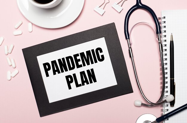 Photo on a light pink background, a notebook with a pen, stethoscope, white pills, paper clips and a sheet of paper with the text pandemic plan. medical concept