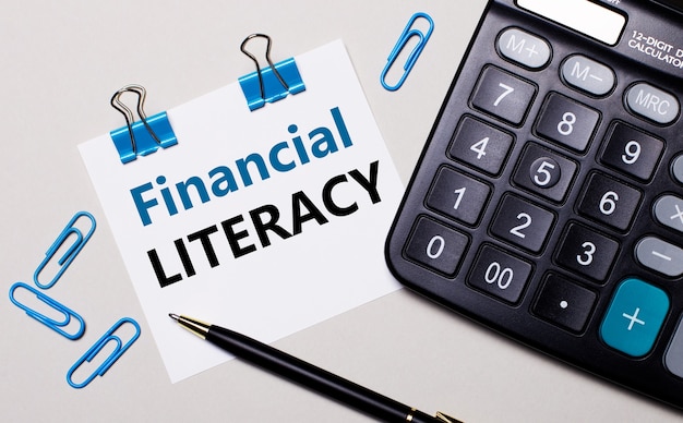 On a light surface, a calculator, a pen, blue paper clips and a sheet of paper with the text FINANCIAL LITERACY. View from above.