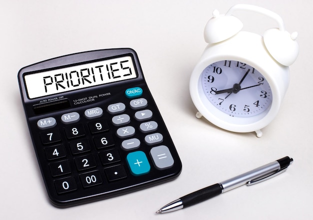 On the light table there is a black calculator with the text PRIORITIES on the scoreboard a pen and a white alarm clock Business concept