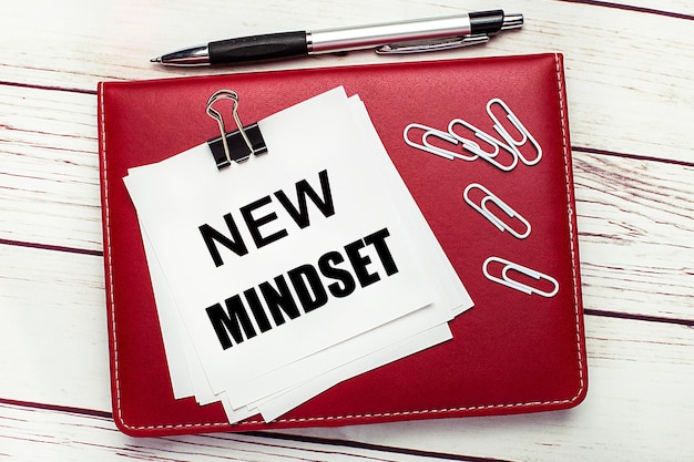 On a light wooden background a burgundy pen and notebook On the notebook has white paper clips and white paper with the text NEW MINDSET Business concept