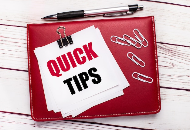 Photo on a light wooden background, a burgundy pen and notebook. on the notebook has white paper clips and white paper with the text quick tips. business concept