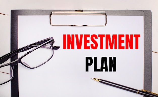 On a light wooden background glasses, a pen and a sheet of paper with the text INVESTMENT PLAN. Business concept