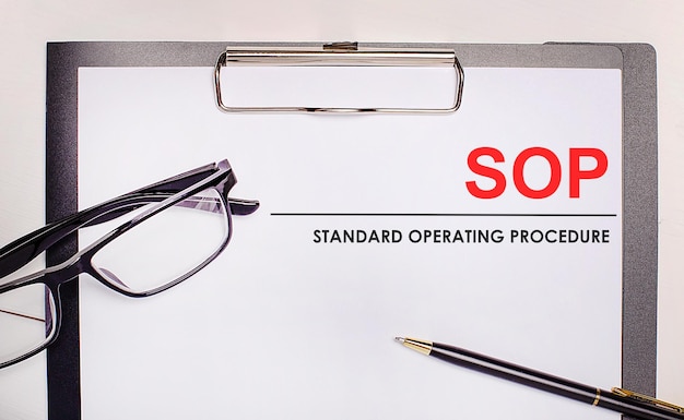 On a light wooden background glasses a pen and a sheet of paper with the text SOP Standard Operating Procedure Business concept