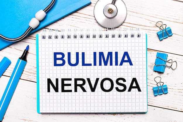 On a light wooden background a stethoscope a blue notepad blue paper clips a blue marker and a sheet of paper with the text BULIMIA NERVOSA Medical concept