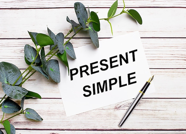 On a light wooden table there is a eucalyptus branch a fountain pen and a sheet of paper with the text PRESENT SIMPLE