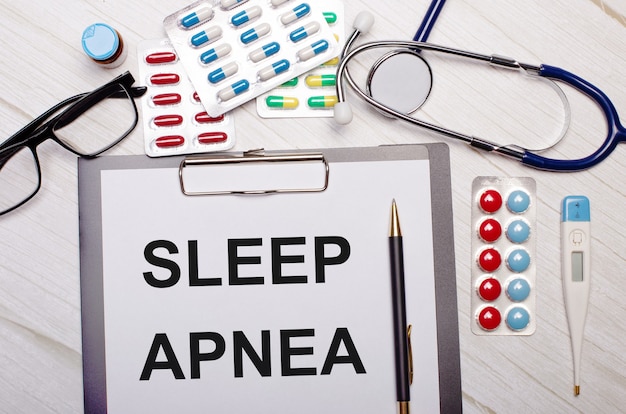 On a light wooden table there is paper with the inscription SLEEP APNEA, a stethoscope, colorful pills, glasses and a pen. Medical concept